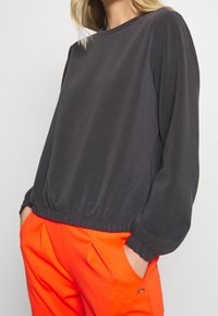 Maglietta a maniche lunghe grigio scuro con scollatura arrotondata e vita elasticizzata, abbinata a pantaloni arancioni brillanti con pince e tasche.