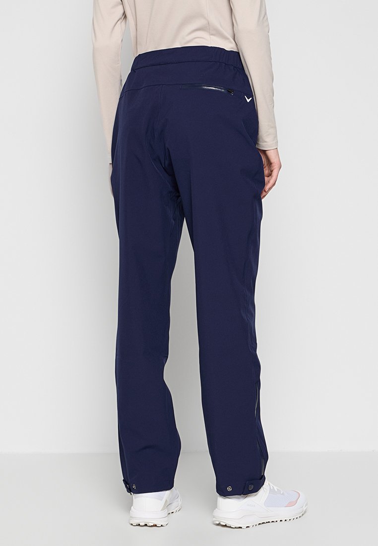 Marineblaue Outdoor-Hosen mit glatter Oberfläche, elastischem Bund, Reißverschlusstasche auf der Rückseite und verstellbaren Bündchen an den Knöcheln, kombiniert mit weißen Schuhen.