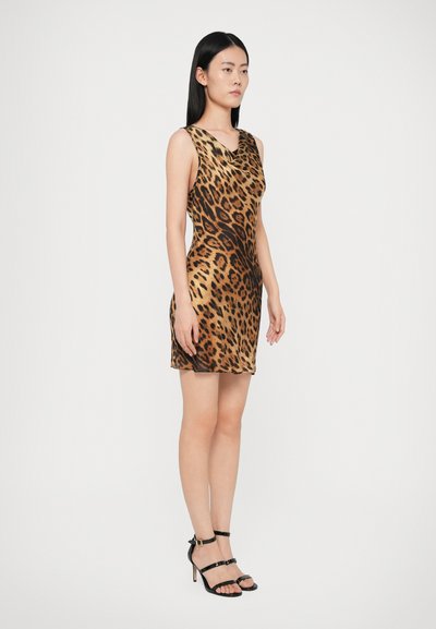 Mulher de pé, vestida com um vestido sem mangas, de comprimento até ao joelho, com estampa de leopardo, e sandálias de salto alto pretas com tiras, contra um fundo branco liso.
