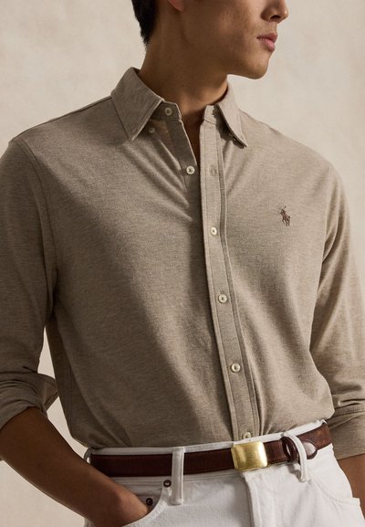 Moški, oblečen v bež srajco z gumbi, zavihanim rokavom, rjav usnjen pas z zlato zaponko in bele hlače, obrnjen na stran.