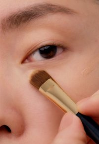 Makeupbørste med et gullfarget metallhåndtak og brune bust som påfører concealer under et mørkebrunt øye på lys hud. Glatt tekstur synlig.