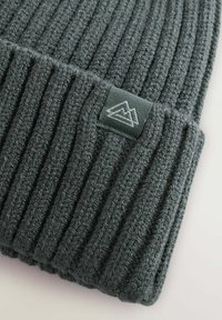 Bonnet en tricot gris foncé avec une texture côtelée, comportant un revers repliable et une étiquette verte avec un logo de montagne.