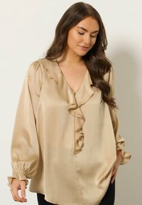 Beżowa satynowa bluzka z dekoltem w serek, falbaniastym przodem i bufkami z elastycznymi mankietami. Gładka tekstura i luźny krój.