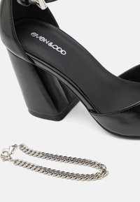 Sort højhælet sko med en glat patentfinish, firkantet tå og kraftig hæl. En sølvfarvet kædeaccent er placeret ved siden af skoen.