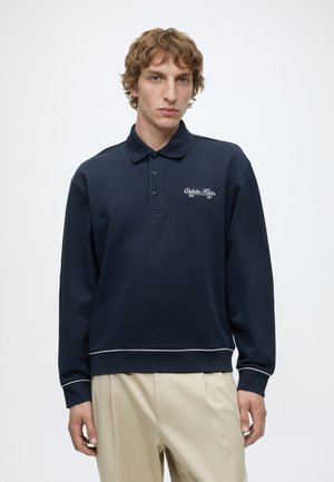 Junger Mann mit welligem Haar, der ein dunkelblaues, langärmeliges Polohemd mit "Calvin Klein"-Logo und beige Hosen trägt.