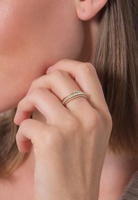 Goldring-Stapeln mit glatten Bändern, eines mit klaren Edelsteinen. Die Handposition nahe dem Gesicht hebt die strukturierte Haut und das Haar hervor.