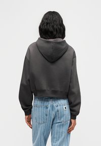 Άτομο με κυματιστά μαλλιά μέχρι τους ώμους, φοράει κοντή σκουρόγριζα μπλούζα με κουκούλα και ψηλόμεσα ριγέ ανοιχτό μπλε τζιν, κοιτάζει προς τα πίσω.