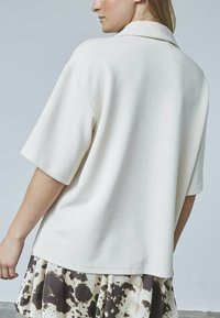 Løs pasform, kortærmet top i blødt cremefarvet stof med ribbet tekstur. Har en krave og sideslidser. Sat sammen med en mønstret nederdel.