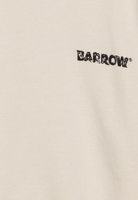 Camiseta de tela color crema con un logo negro de "BARROW" ligeramente desgastado en el pecho, con una textura suave y lisa.