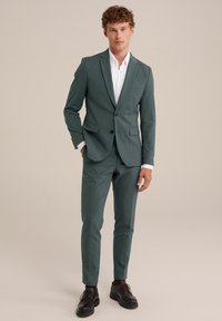 Traje verde entallado con un corte slim, chaqueta de un solo botón y solapas muesca. Combinado con una camiseta blanca y zapatos negros de cordones.