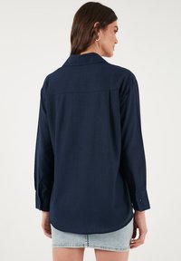 Camicia a maniche lunghe blu navy realizzata in tessuto liscio, con colletto, polsini con bottoni e yoke posteriore con pence per un ulteriore dettaglio.