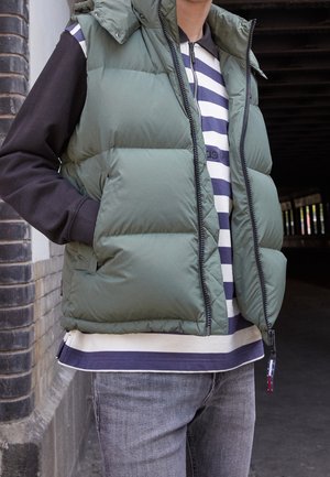 Osoba ubrana w zieloną puchową kamizelkę na czarnej bluzie z kapturem i granatową koszulę w białe paski, stojąca obok muru z cegieł w zacienionym miejscu na zewnątrz.