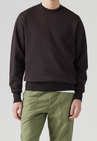 Uomo che indossa una felpa nera a maniche lunghe e pantaloni verde oliva, in piedi contro uno sfondo grigio chiaro uniforme.