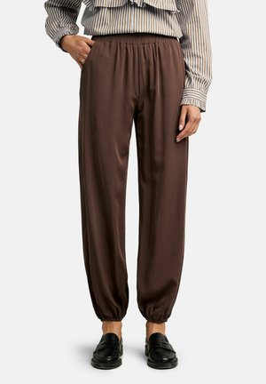 Femme portant un pantalon ample marron à poignets élastiques, des mocassins noirs et un chemisier beige et blanc à rayures avec des détails à volants.
