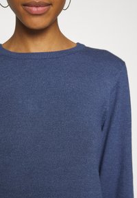 Modrý pletený svetr s kulatým výstřihem, měkkou texturou a dlouhými rukávy. Zobrazuje detailní pohled na rovnoměrný pletený vzor látky.