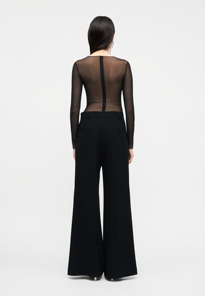 Γυναίκα με μακριά σκούρα μαλλιά, που φοράει διαφανές μαύρο τοπ με μακριά μανίκια και ψηλόμεσα, φαρδιά μαύρα παντελόνια, στέκεται με την πλάτη προς το μέρος μας.