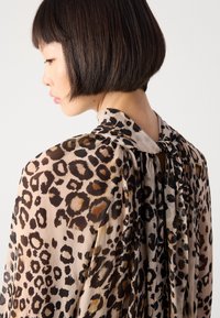 Femme avec un carré court portant un chemisier en tissu transparent à motifs léopard noué dans le dos, faisant face à gauche sur fond uni.