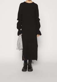 Svart stickad klänning med långa ärmar och ribbad textur; sidospalt. Kombinerad med svarta snörstövlar, håller en silverfärgad fransväska.