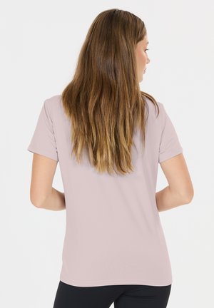 Woman with long brown hair wearing a light pink short-sleeve shirt and black pants, facing away against a plain white background.