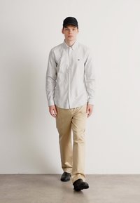 Wit en blauwe gestreepte overhemd, beige broek en zwarte pet. Model staat tegen een eenvoudige achtergrond en toont casual kleding.