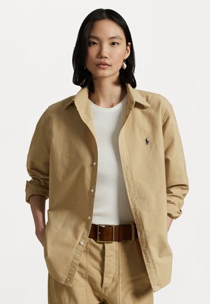 Camicia beige a bottoni con una vestibilità rilassata, realizzata in cotone. Presenta un piccolo logo sul petto ed è abbinata a un top bianco a coste e pantaloni color beige.