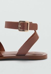 Ruskeat litteät sandaalit nilkkahihnalla ja kultaisella neliösoljella, joissa on kaksi kulmaista etuhihnaa yksinkertaisella taustalla.