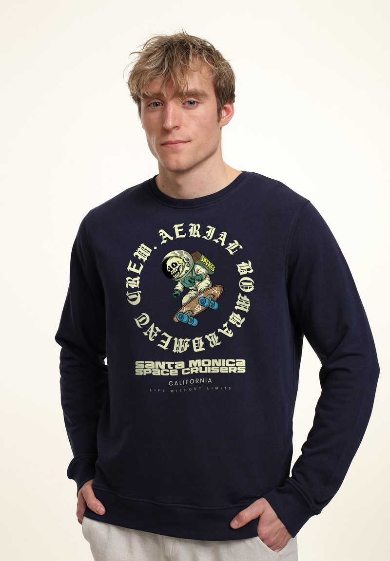Sudadera azul marino de manga larga, con un gráfico de un esqueleto en un traje de astronauta sobre una patineta. El texto incluye "Santa Monica Space Cruisers."