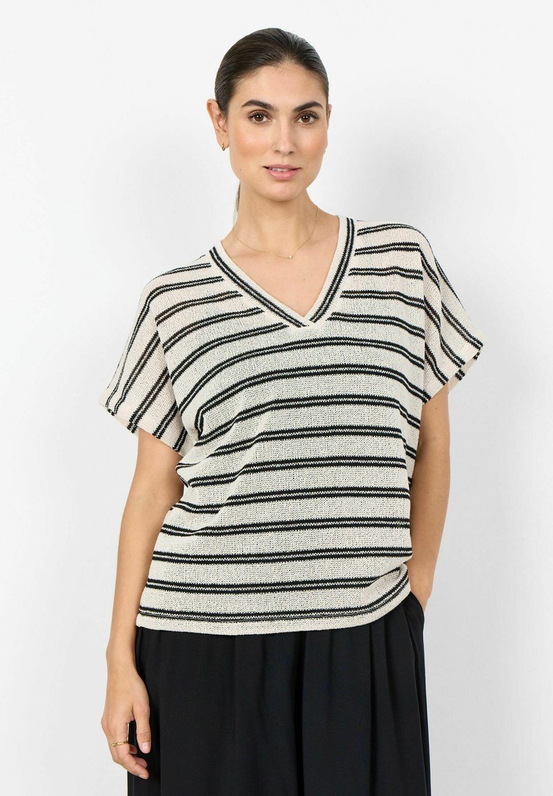 Femme portant un haut en tricot blanc à manches courtes avec des rayures horizontales noires et un col en V, associé à un pantalon noir ample, debout devant un fond blanc.