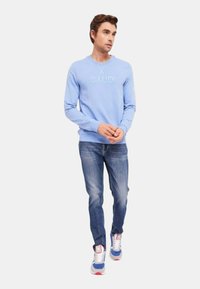 Felpa azzurra a girocollo con logo ricamato "GAUDI", abbinata a jeans blu e sneakers multicolori. Vestibilità e design casual.