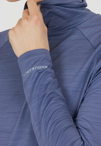 Langärmliges, hellblaues Sportoberteil mit hohem Kragen, strukturiertem Stoff und dezenten horizontalen Streifen. Logo "ENDURANCE" auf dem Ärmel.