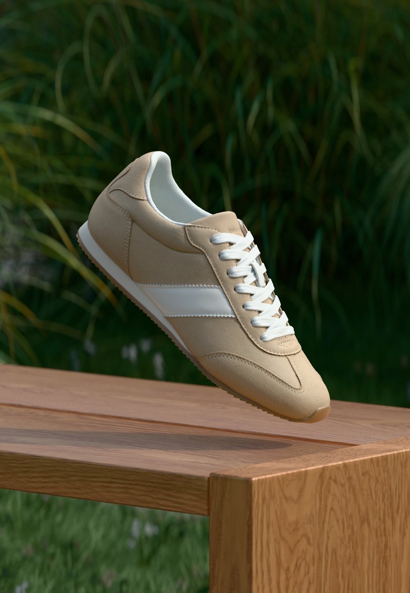 Chaussure de sport beige avec une finition en daim lisse, des lacets blancs et une bande blanche contrastante. Design minimaliste avec un bout arrondi et une semelle texturée.