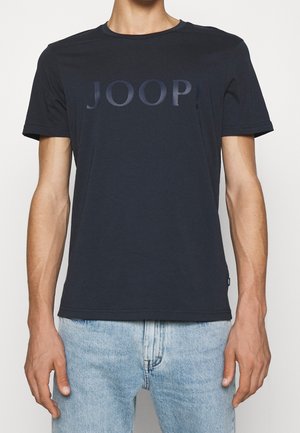 Mężczyzna w czarnej koszulce z logo JOOP i jasnoniebieskich dżinsach, stojący na jednolitym jasnym tle.