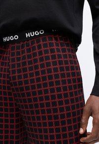 Schwarze Jogginghosen mit wiederholtem roten "HUGO"-Muster, ausgestattet mit einem schwarzen Bund mit "HUGO"-Logo. Glatte Textur, figurschnitt.