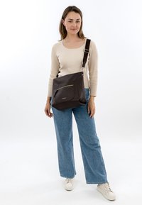 Braune Schultertasche aus strukturiertem Leder mit einer vorderen Reißverschlusstasche und verstellbarem Riemen. Das Modell trägt hellblaue Jeans und weiße Turnschuhe.