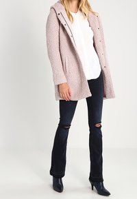 Lichtroze, gestructureerde jas met een capuchon, drukknoopsluitingen en zijzakken. Gepaard met donkere, flared jeans met kniebescheuringen en zwarte enkellaarzen.