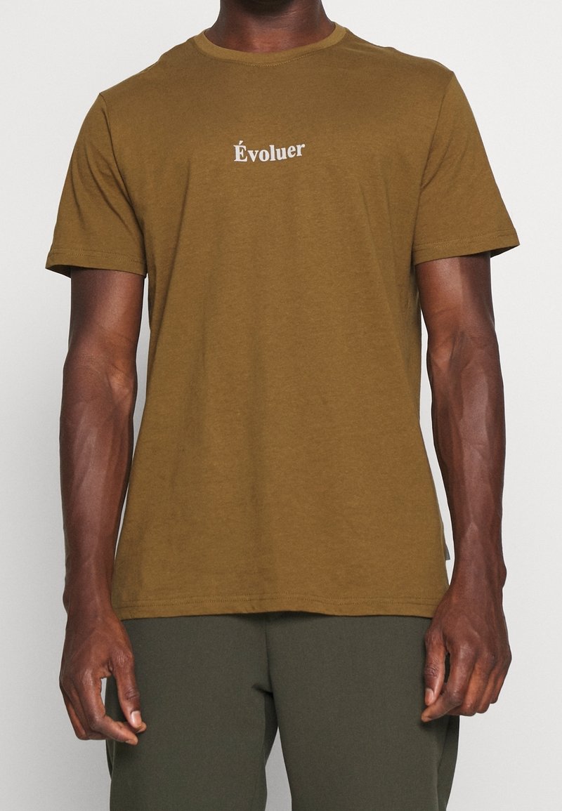 Homme portant un t-shirt à manches courtes vert olive avec le mot "Évoluer" imprimé en blanc sur la poitrine.