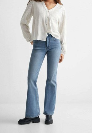 Kvinde iført hvid langærmet skjorte med knapper, lyseblå højtaljede viddejeans og sorte chunky støvler stående på hvidt gulv.