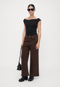 Vrouw in een zwarte off-shoulder top, bruine wijde leopardprint broek, zwarte zonnebril, met een zwarte handtas en zwarte platformlaarzen.