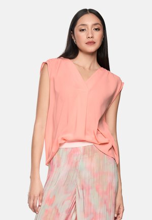 Woman with long dark hair wearing a loose coral sleeveless top and pleated pastel multicolor skirt against a plain background.