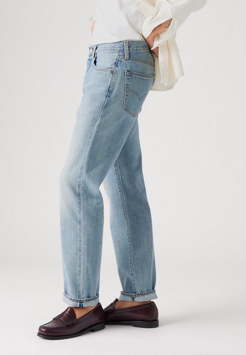 Jeans de pierna recta azul claro con un acabado desgastado, cinco bolsillos y dobladillo enrollado, combinados con mocasines marrón oscuro y una camiseta blanca.
