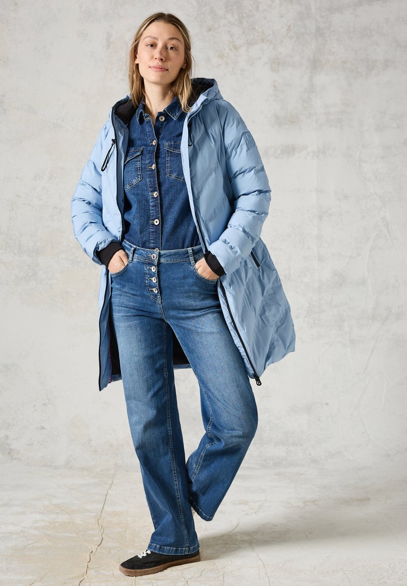 Ljusblå puffjacka med huva, bärs över en denimskjorta och utsvängda jeans, med silverknappar och svarta accenter på ärmarna.