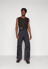 Czarne spodnie techniczne z dwiema kieszeniami na zamek, regulowanym pasem, w połączeniu z bezrękawną ciemną bluzką. Odbijający logo detal.