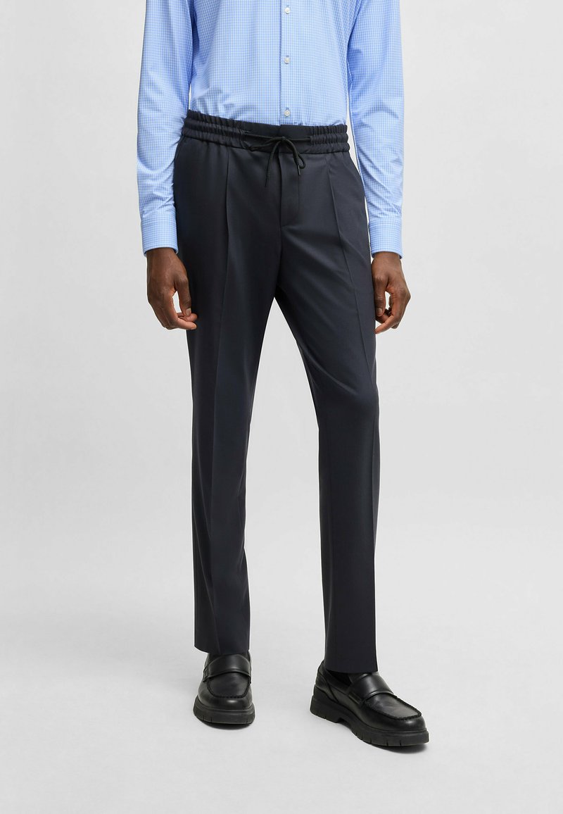 Dark navy trousers with an elastic waistband, drawstring, and smooth fabric. The model wears a light blue checked shirt and black shoes.
