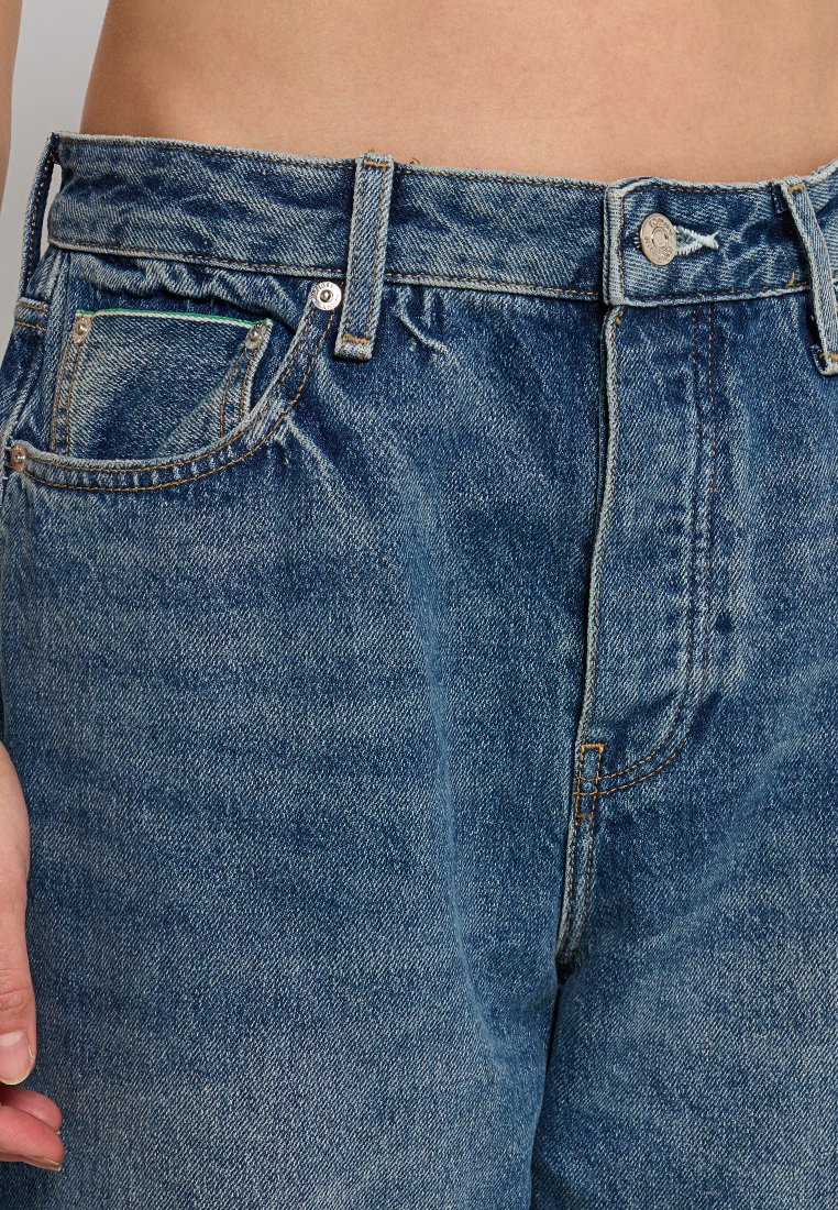 Jeans denim w średnim niebieskim kolorze z wysokim stanem, z pięcioma kieszeniami, metalowym zapięciem i widocznymi detalami szwów.