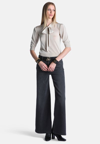 Jeune femme portant un haut beige à manches courtes avec un noeud papillon, un pantalon noir à jambes larges, des bottes noires et des bracelets de poignet et une ceinture noirs assortis.