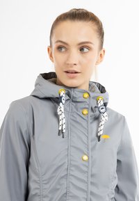 Jachetă gri impermeabilă cu glugă, având șnururi decorative albe și bleumarin, feronerie galbenă și un mic logo cu umbrelă galbenă.