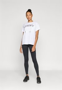 Λευκό γραφικό μπλουζάκι με την επιγραφή "DKNY 1988 NEW YORK" σε ασημί τύπωμα, συνδυασμένο με μαύρα κολάν και μαύρα αθλητικά παπούτσια. Ομαλό ύφασμα, άνετη γραμμή.