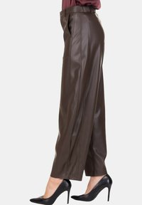 Donna con pantaloni di pelle marroni a vita alta e tacchi alti neri a punta, in piedi di lato contro uno sfondo semplice.