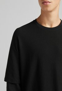 Černé tričko s krátkým rukávem vyrobené z texturované žebrované látky s kulatým výstřihem. Disponuje volným střihem a subtilními detaily šití.