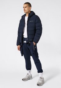 Granatowa kurtka puchowa z suwakiem z przodu, z kapturem i logo na klatce piersiowej. Zestawiona z granatowymi dresami i szarymi trampkami.